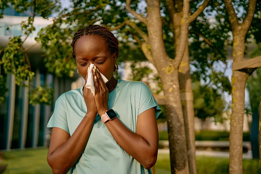 A woman standing outdoors near trees blows her nose into a tissue, suggesting seasonal allergy symptoms. She wears a teal t-shirt and a smartwatch.